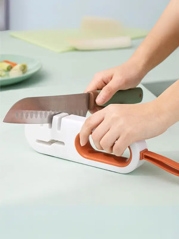 A person's hand using a pull-through knife sharpener on a kitchen counter. The sharpener has two grinding stages and a compartment for sharpening scissors